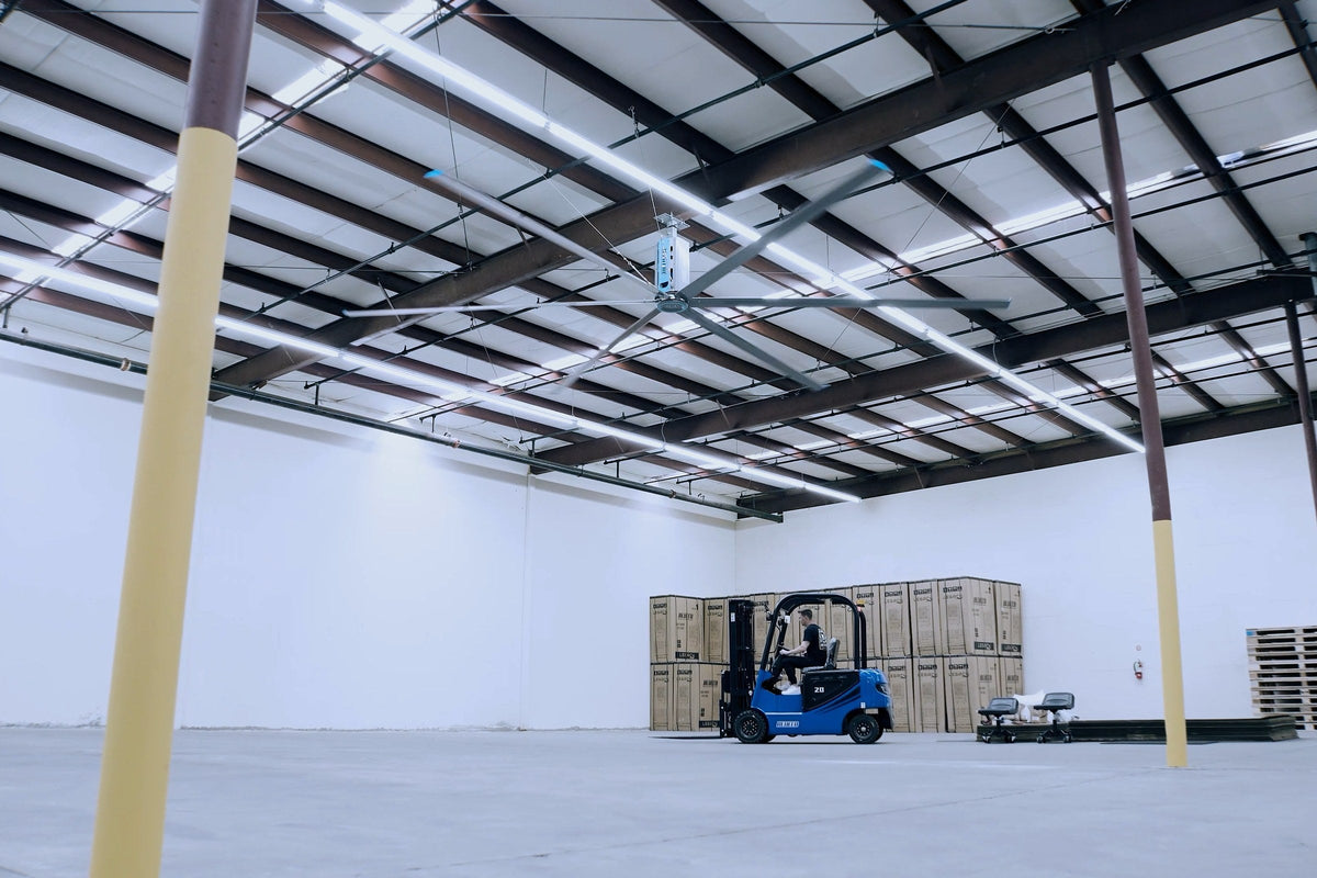 Large warehouse bay under HVLS fans, a BLUECO electric forklift by stacked cartons
