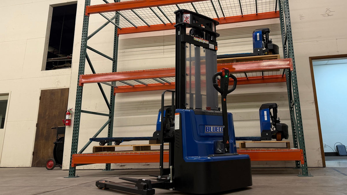 BLUECO straddle-leg stacker in front of orange warehouse racking with two electric pallet jacks on the shelves