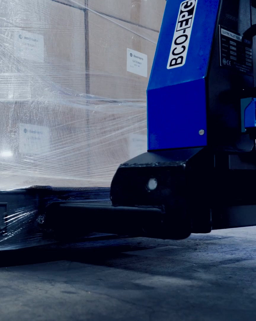 Electric pallet jack forks under a shrink-wrapped pallet, seen low from the side
