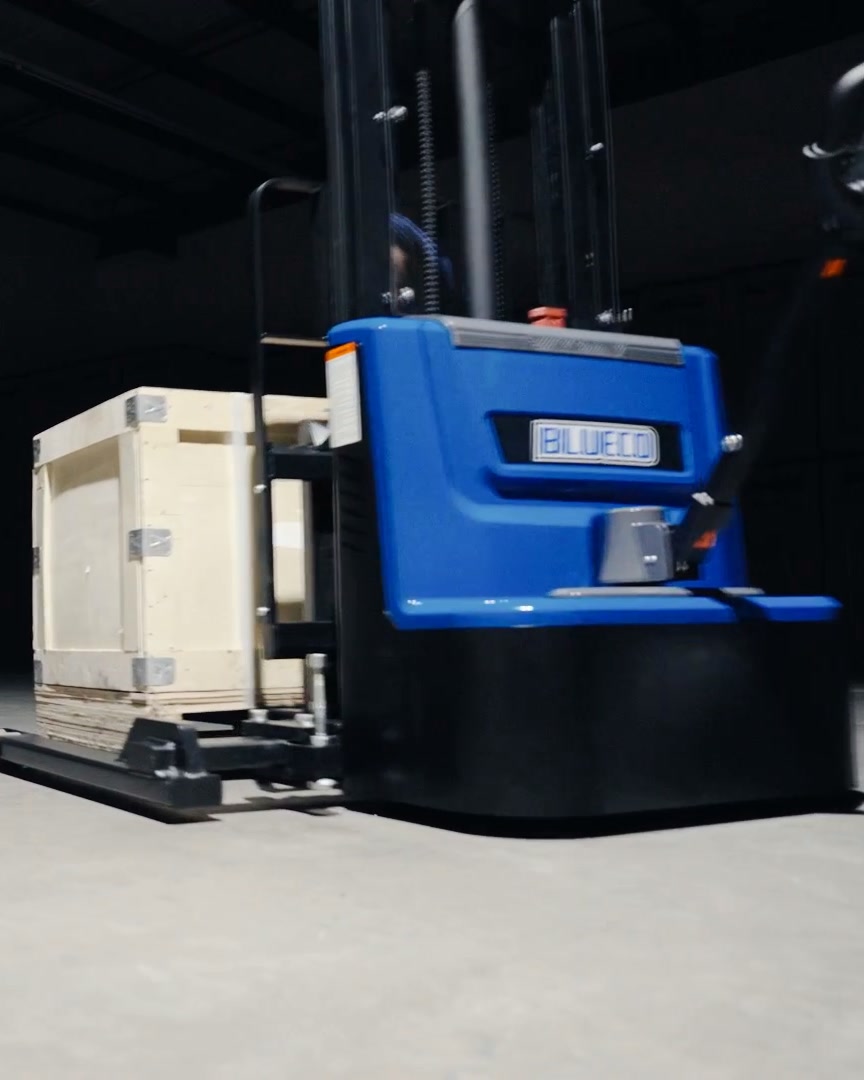 BLUECO straddle-leg stacker carrying a wooden crate across a dark studio floor