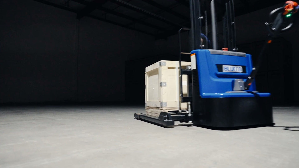 BLUECO straddle-leg stacker carrying a wooden crate across a dark studio floor